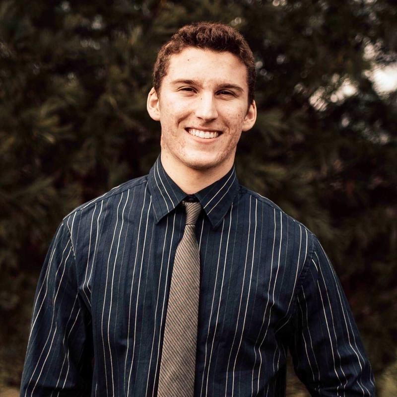 Samuel Atkinson stands smiling against a tree backdrop